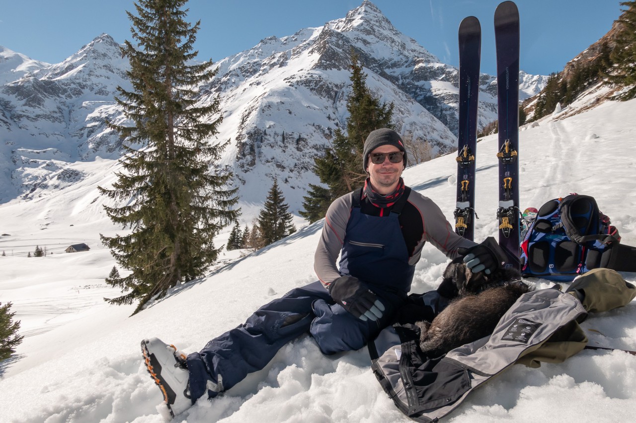 Man sitting in the snow with his dog in the mountains with his skis set up behind him in the snow