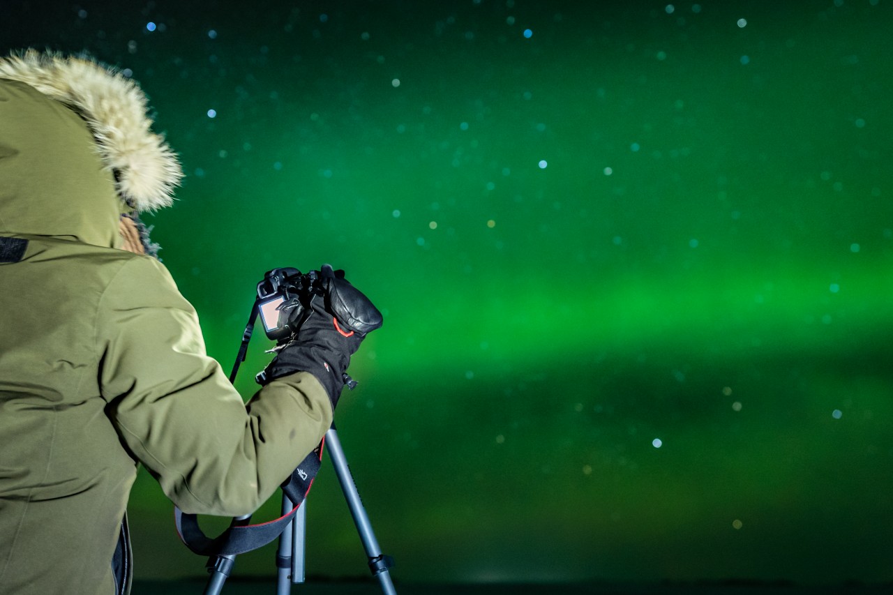 Persona con spessa giacca invernale e guanti, macchina fotografica e treppiede fotografa il verde cielo notturno